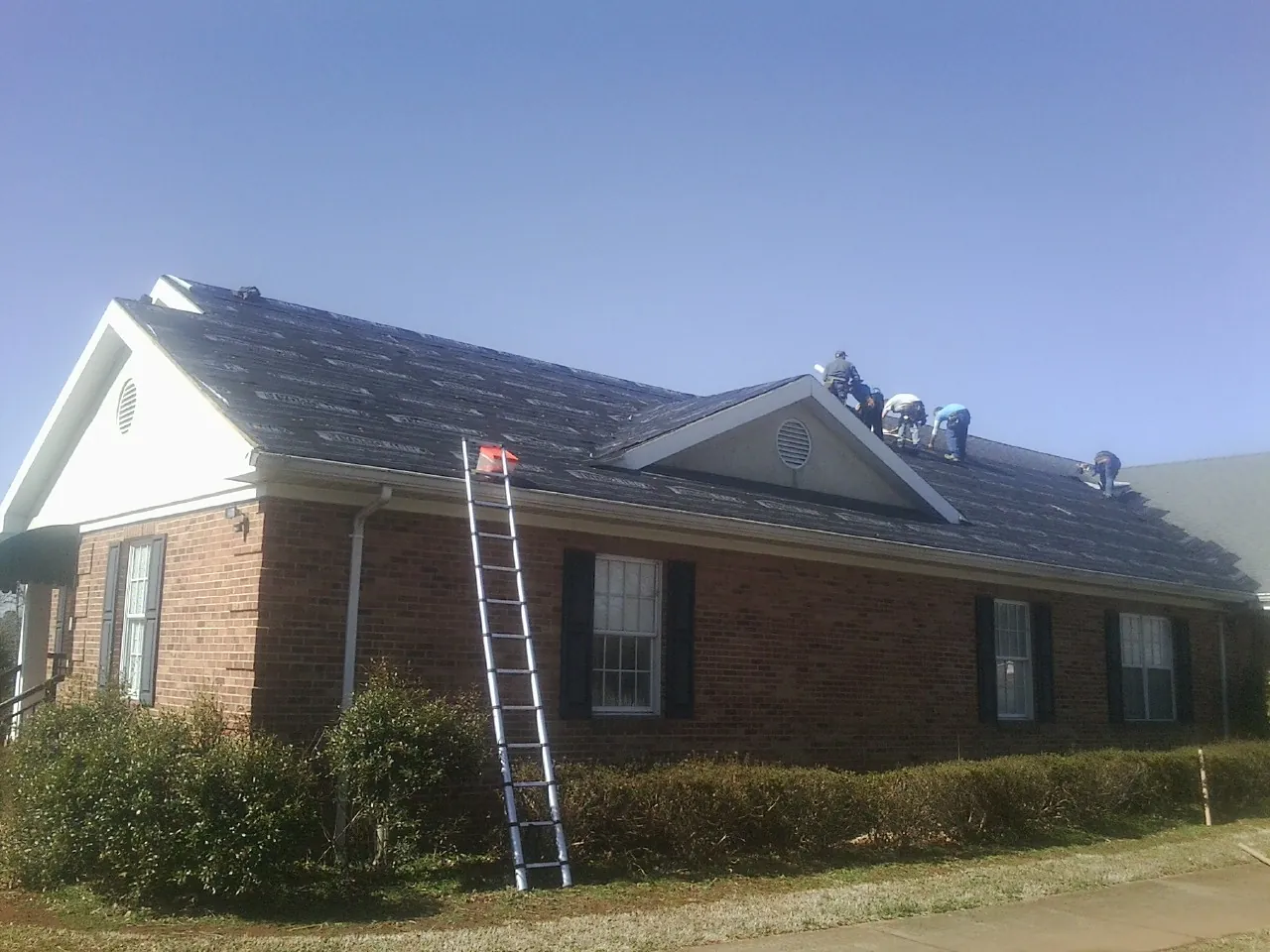 Crew tearing shingles off a brick ranch roof with a ladder set — Metal Roof Repair work by our crew in Borup