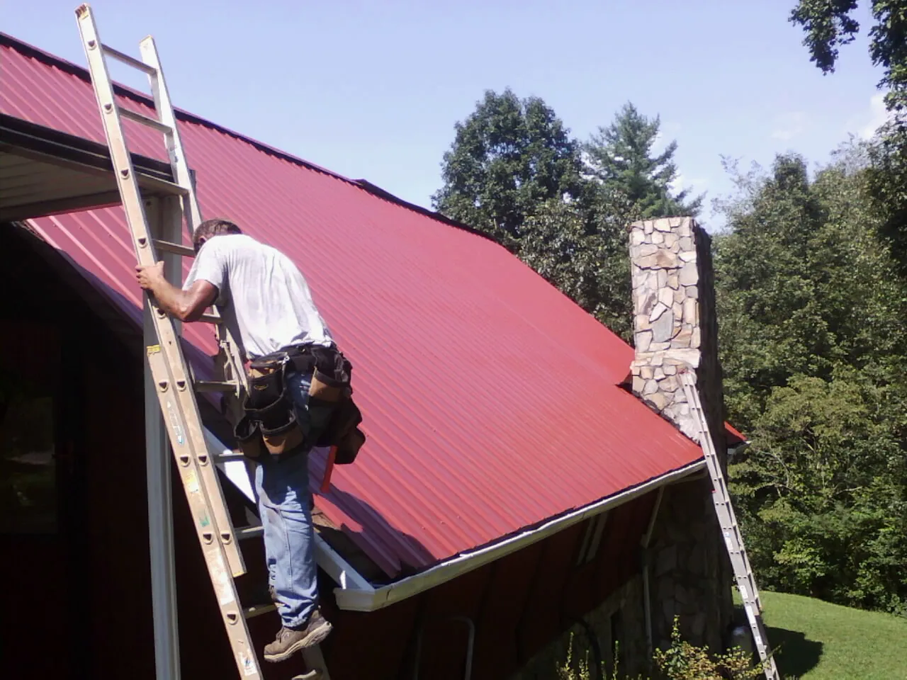 Roofer on a ladder beside a red metal roof and stone chimney — Soffit & Fascia Repair by Roofing Brothers in Borup
